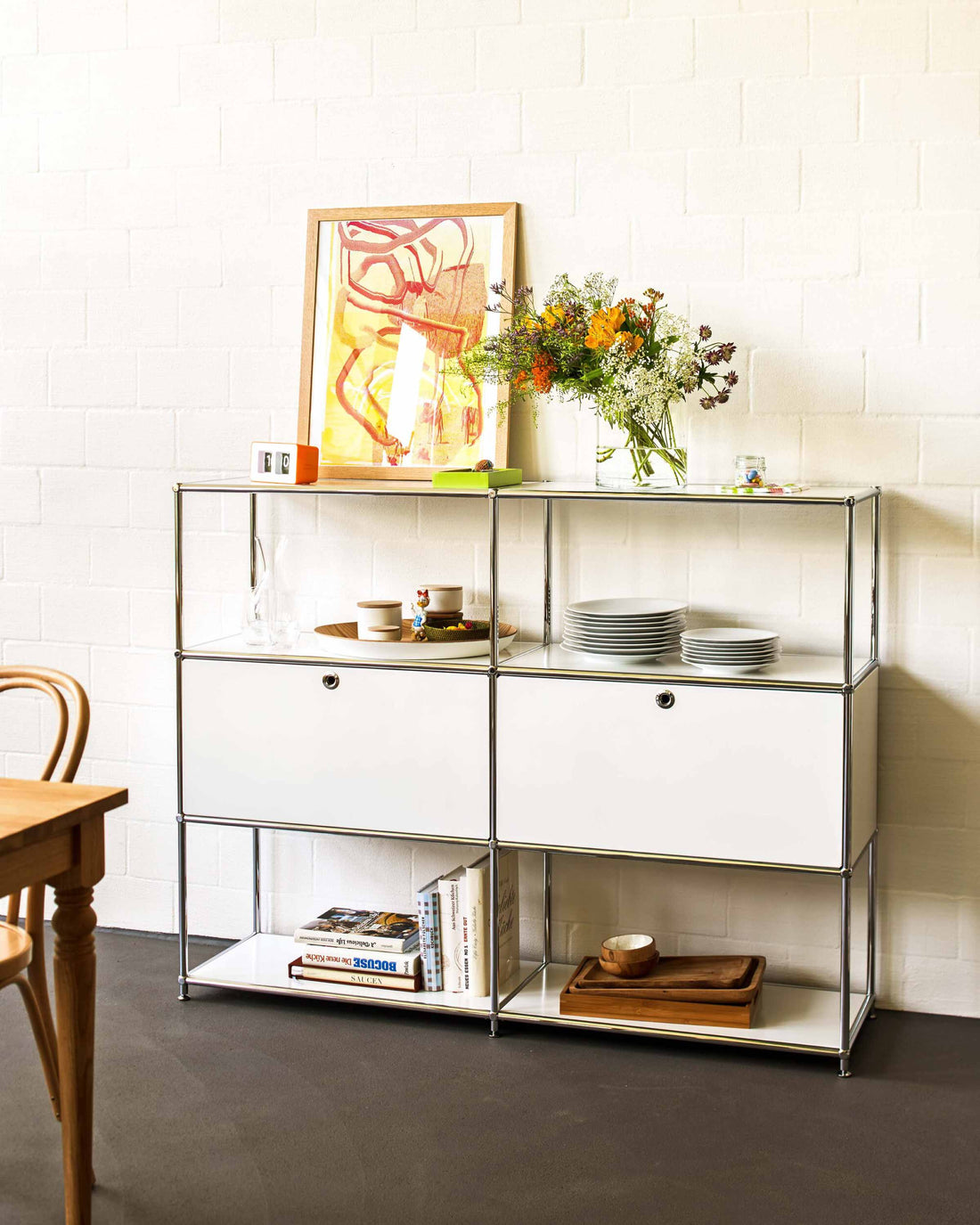 Weisses FLEXCUBE‑Regal mit Geschirr, Büchern, einer Vase mit Blumen und einem gerahmten Kunstbild vor einer hellen Wand.