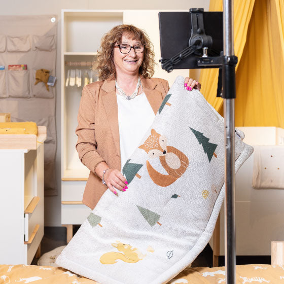 Person präsentiert eine Kinderdecke mit Tiermotiven vor einer Kameraeinrichtung in einem Kinderzimmer‑Showroom.